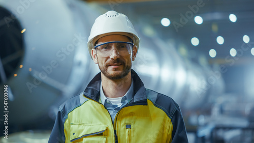 Portrait of Smiling Professional Heavy Industry Engineer / Worker Wearing Safety Uniform, Goggles and Hard Hat. In the Background Unfocused Large Industrial Factory where Welding Sparks Flying