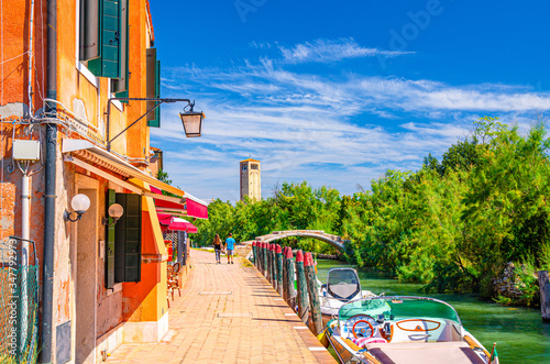 Torcello island with embankment along water canal with motor boats and buildings along promenade. Stone Devil bridge, green trees and tower background. Venetian Lagoon, Veneto Region, Italy.
