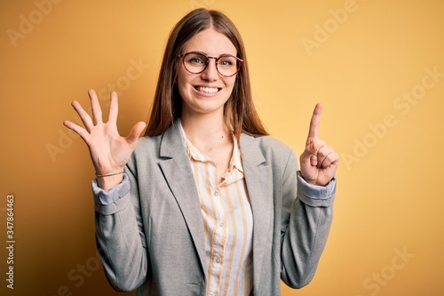 Young beautiful redhead woman wearing jacket and glasses over isolated yellow background showing and pointing up with fingers number six while smiling confident and happy.