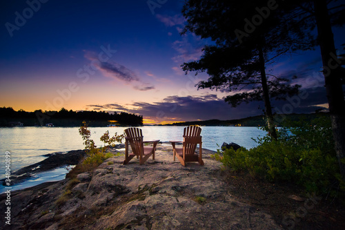 Two Muskoka chairs sitting on a rock formation facing a calm lake at dusk in cottage country. 