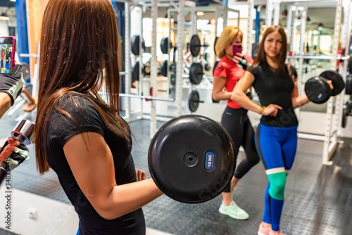 Two young attractive fitness girls take photo with dumbbell in gym mirror