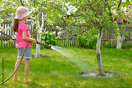 Little girl in a hat is watering a tree with a hose at the dacha.