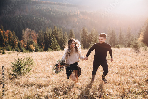 Stylish model couple in the autumn mountains. A young guy and a girl run along the slope against the background of the forest and mountain peaks at sunset. Girl holds in her hands a bouquet of flowers