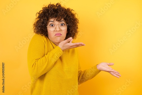 Beautiful Arab businesswoman pointing aside with both hands showing something strange and saying: I don't know what is this. Standing against yellow background. Advertisement concept.