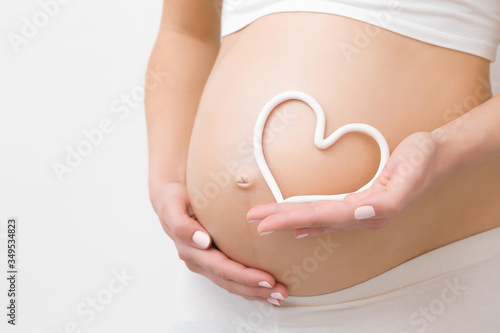 Young woman hand holding white heart shape on naked belly. Emotional loving moment in pregnancy time - 30 weeks. Baby expectation. Love, happiness and safety concept. Closeup.