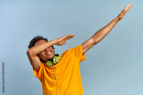 Cheerful African-American young guy joyfully raises hands up covering face as dub dancing sign posing on blue background with headphones in yellow t-shirt. Сoncept of music subscription, online radio