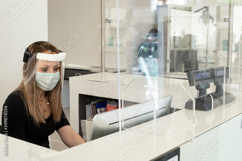 segretaria di un locale nella postazione di lavoro alle prese con il telefono e il lavoro al computer equipaggiata di maschera professionale e protetta da un plexiglas contro 