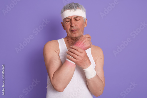 Horizontal shot of senior man dresses white sleeveless t shirt, hurts his wrist during sport training, white haired male posing isolated over lilac studio background. Fitness, health care concept.