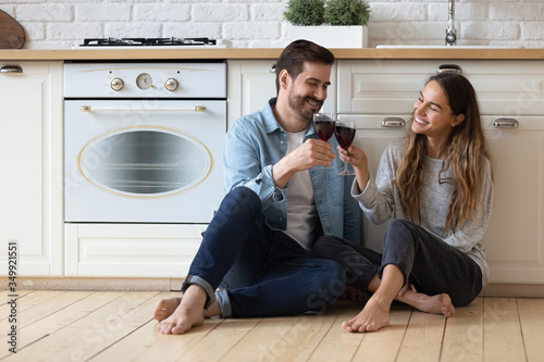 Happy young husband and wife sit relax on wooden floor in modern design kitchen enjoy family leisure weekend together, overjoyed millennial couple having romantic date celebrate wedding anniversary