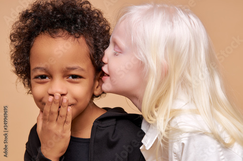 albino girl with pale skin and white hair color tells a secret in the ear of multiracial african boy. cute boy with black skin attentively listen to secret. isolated