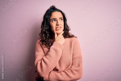 Young beautiful woman with curly hair wearing casual sweater over isolated pink background Thinking worried about a question, concerned and nervous with hand on chin