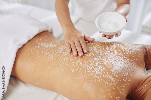 Beautician exfoliating back of young woman with salt scrub before massage