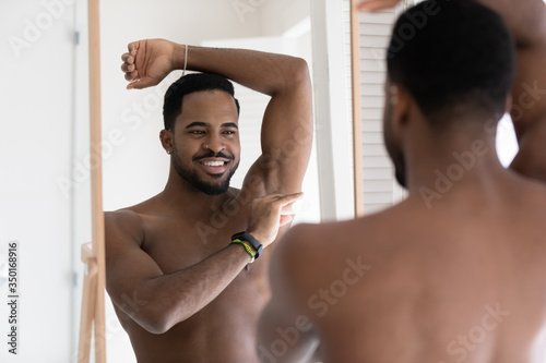 Smiling young naked african American man look in mirror in bathroom after shower apply deodorant on armpit, happy handsome millennial male use dry stick antiperspirant in bath, body hygiene concept