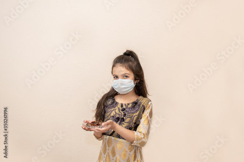 Small girl wearing traditional pakistani dress and mask and celebrating Ramadan Kareem. Holds plate of dates.