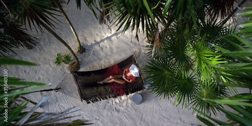 Woman rests in loose red summer dress and hat hanging in hammock surrounded by palm trees at sunset
