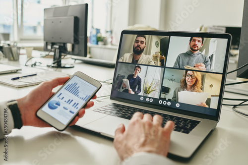 Close-up of businessman working in empty office and using smartphone to view graph while discussing sales with colleagues via video conferencing app