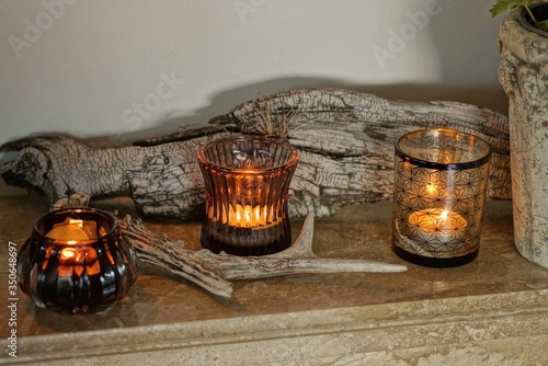 three burning yellow candles in brown glasses stand on a table against a gray wall in the room