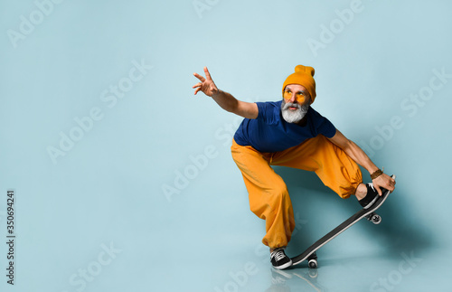 Gray-bearded grandpa in t-shirt, sunglasses, orange pants, hat, gumshoes. Riding black skateboard, posing on blue background