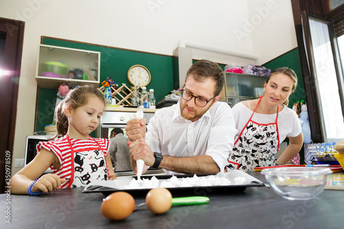 famigliola felice prepara un dolce nella cucina dove la mamma con la figlia guardano con simpatica diffidenza i movimenti del marito