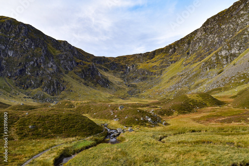 Corrie Fee in Glen Clova, Angus Glens, Scottish Highlands with pathway leading to mountains, waterfall in distance. Blue sky and clouds, sunny day.