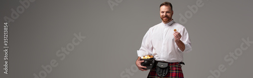 smiling Scottish redhead man in red kilt holding potty with gold coins on grey background, panoramic crop