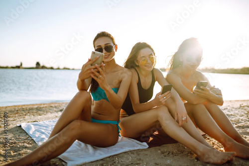 Three beautiful girls using mobile smart phone at the beach. Girls sunbathing on the beach. Young girls doing selfie with phone. Enjoy summer time.