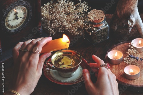 Wiccan witch wearing vintage jewelry holding yellow candle and pouring wax into a red gold vintage teacup as a divination. Reading Candle Wax - Carromancy, Ceroscopy among nature items dried flowers