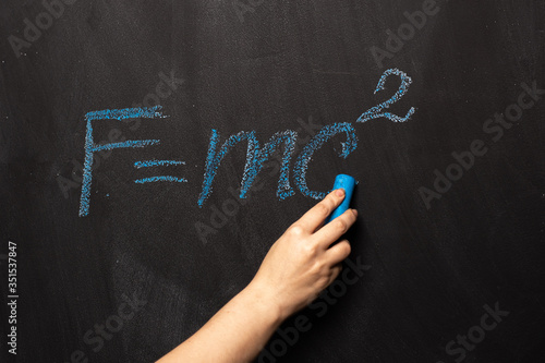 The physical formula of Einstein's theory is written in blue chalk on a blackboard. FMC2 is written by a science teacher or student in the classroom.