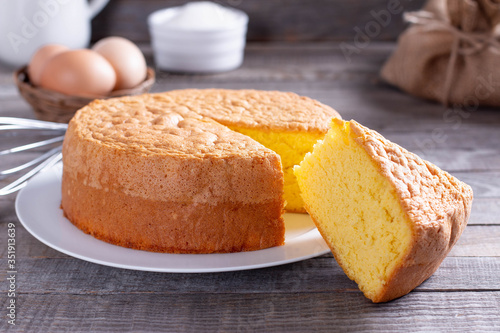 Homemade round sponge cake or chiffon cake on white plate so soft and delicious with ingredients: eggs, flour, milk on wood table