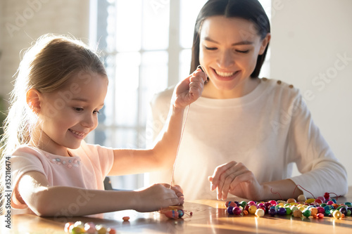 Loving young mother teach excited little daughter string thread wooden beads making stylish bracelets, happy mom or nanny and small preschooler girl child engaged in creative activity or game at home