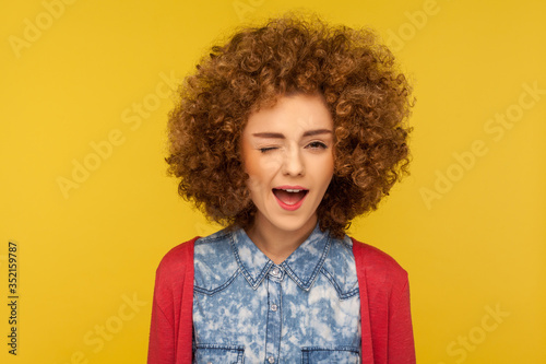 Closeup portrait of playful happy charming woman with curly hair in casual outfit blinking eye as having cunning idea, winking and flirting to camera. indoor studio shot isolated on yellow background