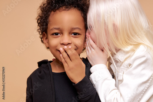 portrait of sweet children having secret isolated in studio, beautiful kids of diverse nationalities stand together, albino girl with blonde hair tells secret in multiracial black boy's ear
