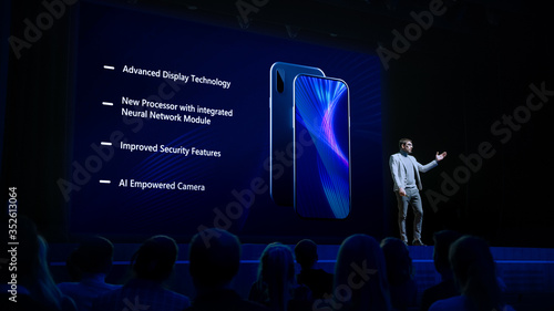 Live Event with Brand New Products Reveal: Keynote Speaker Presents Smartphone Device to Audience. Movie Theater Screen Shows Mock-up Touch Screen Phone with High-Tech Features and Top Highlights