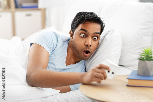 people, bedtime and rest concept - overslept indian man looking at alarm clock lying in bed at home