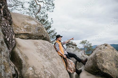 A young man in a hat sits on top of a cliff and looks around. Travel concept