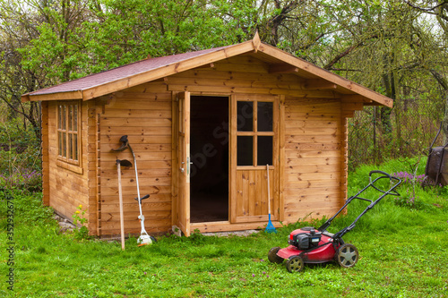 Garden shed with hoe, string trimmer, rake and grass-cutter. Gardening tools shed. Garden house on lawn in garden. Wooden tool-shed. Hovel made of timber in domestic environment.