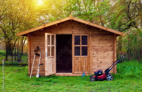 Hovel after work in evening, golden hour. Garden shed (front view) with hoe, string trimmer, rake and grass-cutter. Gardening tools shed. Garden house on lawn in the sunset. Wooden tool-shed.