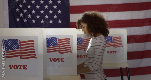 Profile, medium shot, young Hispanic woman in polling station, voting in a booth with US flag in background.