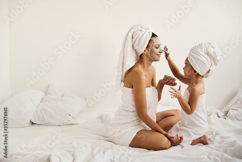 A young caucasian mother and little daughter with wrapped hair in white bath towels apply a clay mask on the mother faces
