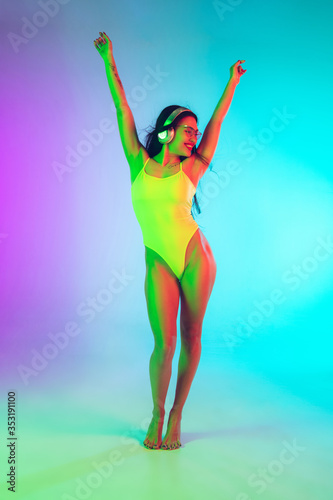 Beautiful young girl dancing with wireless headphones on gradient studio background in neon light. Woman in fashionable bodysuit. Facial expression, summer, weekend, beauty, resort concept. Vacations.