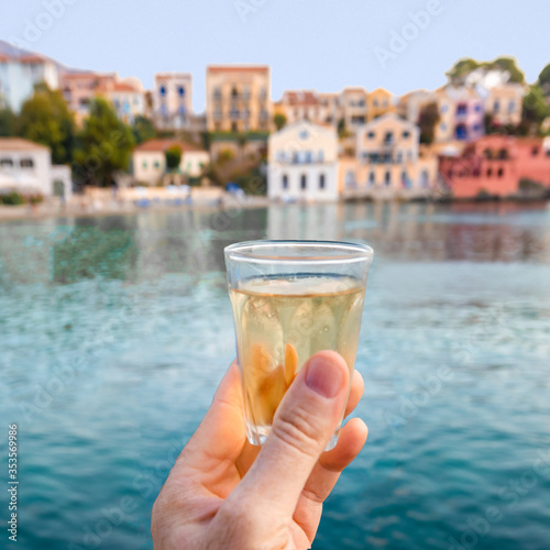 Raising hand holding a glass of retsina white wine in front of the sea and the famous village Assos in Kefallonia island in Greece. Happy holidays toast.