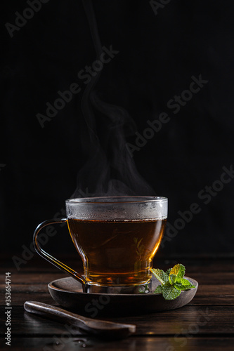 Steaming peppermint infusion in crystal glass with fresh mint leaves, on dark rustic wooden base.