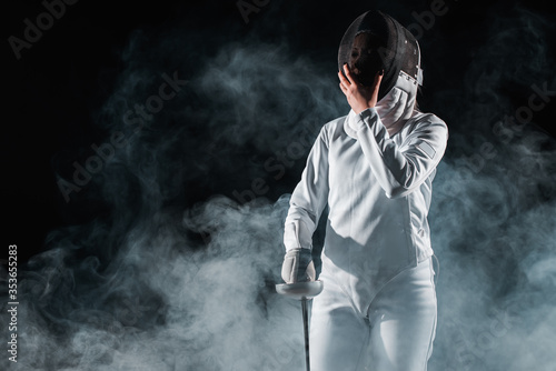 Fencer holding fencing mask and rapier on black background with smoke