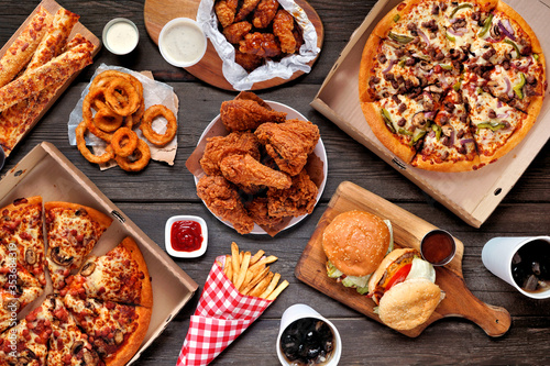 Buffet table scene of take out or delivery foods. Pizza, hamburgers, fried chicken and sides. Above view on a dark wood background.