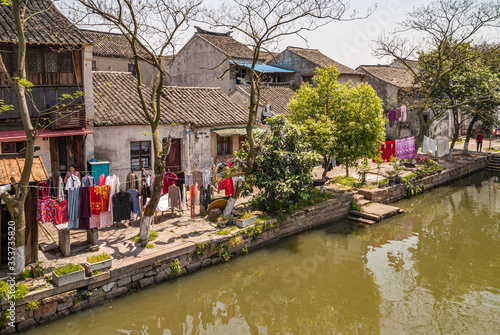 Tongli, JIangsu, China - May 3, 2010: Closeup of lines of drying colorful laundry along green water canal under light blue sky with green foliage and dark roofed dirty white houses.