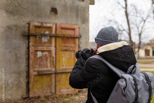 Man in warm jacket and cap with back pack standing back to camera and taking photographs of abandoned building outdoors.