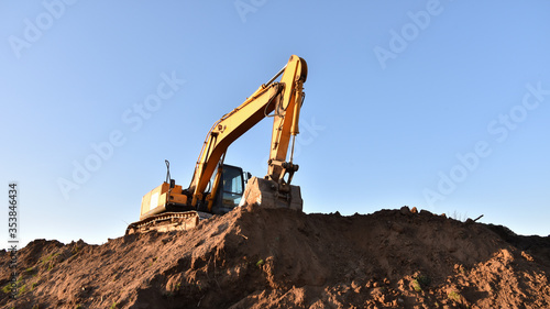 Excavator working at construction site. Backhoe digs ground in sand quarry on blue sky background. Construction machinery for excavation, loading, lifting and hauling of cargo on job sites