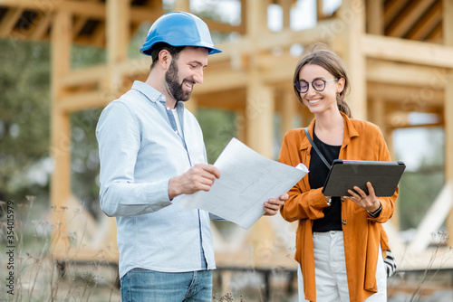 Builder with a female client near the wooden house structure on the construction site outdoors. Building and designing wooden frame house concept