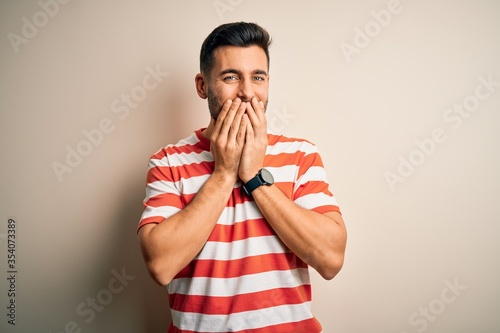 Young handsome man wearing casual striped t-shirt standing over isolated white background laughing and embarrassed giggle covering mouth with hands, gossip and scandal concept