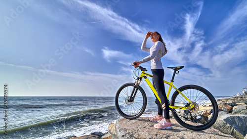 Fitness woman with a bicycle stands on the stones on the seashore and looks into the distance on a clear sunny day. Athletic healthy people with sporty and active lifestyle